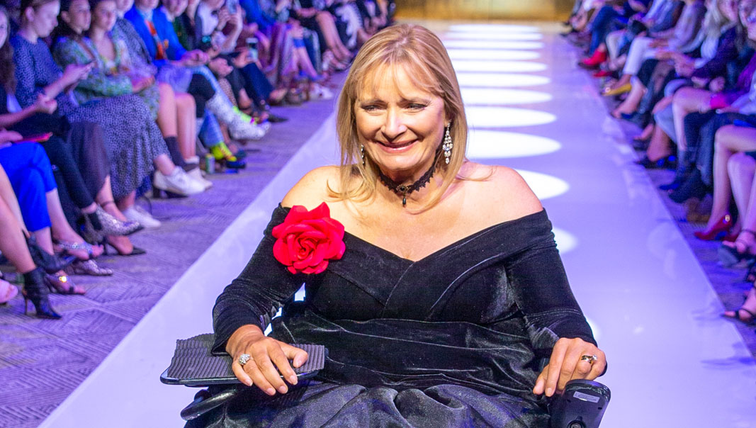 Carol Taylor on the runway at MBFW, woman in a black off-the-shoulder dress in a wheelchair smiling