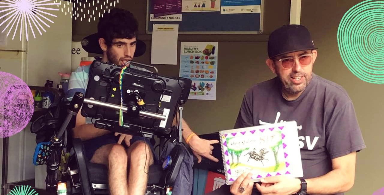 Dale seated in his wheelchair with communication board in front of him, gesturing to a story book being held by his support worker