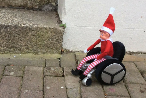Elf on the shelf toy in a wheelchair, outdoors, paved ground