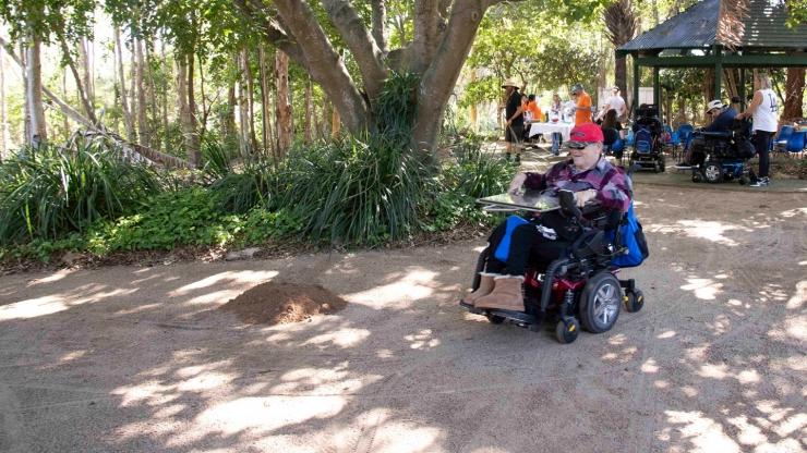 Man in a wheelchair on a garden path, trees, gazebo and other people in the background