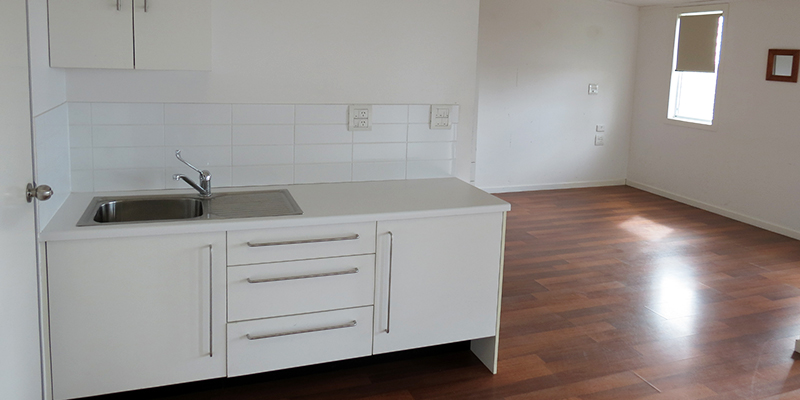 Vacant room with polished wooden floorboards and white kitchenette with sink and cupboards