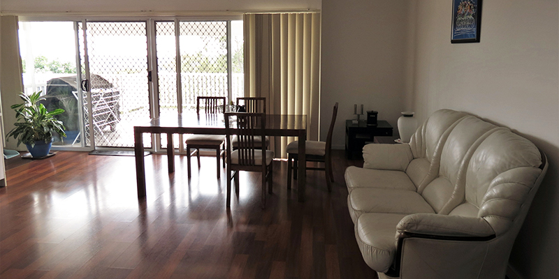 Large living room with polished wooden floorboards. Grey leather couch and dining setting, with sliding doors to deck on back wall.