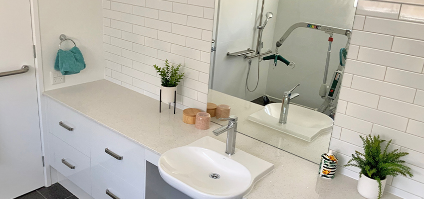 Bathroom vanity with white cabinets and white tiles. Reflected in the mirror are grab bars and a hoist