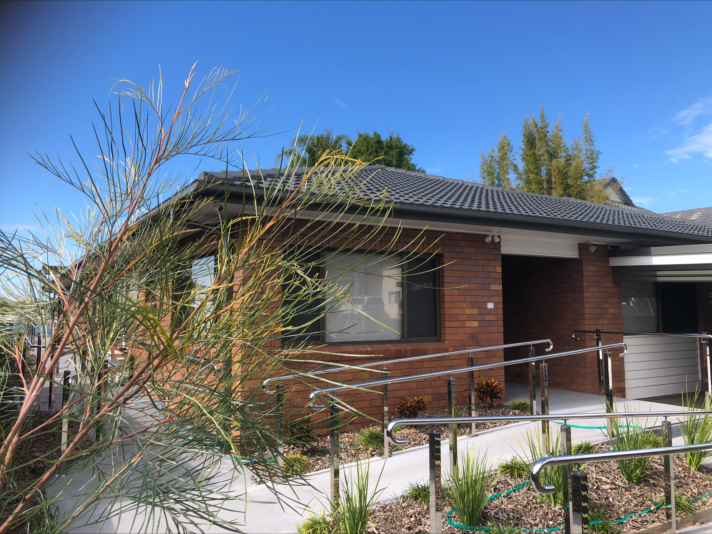 An image showing the front of the property, with a blue sky, green leaves in the foreground and an accessible ramp.