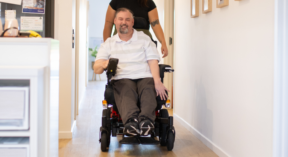 Image showing a man in a wheelchair in a supported independent living arrangement smiling towards the camera, with a support worker in the background