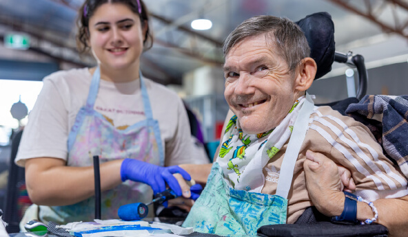 An older man sits in a wheelchair at a table inside of an art studio. He is wearing an art apron and being assisted by a support worker to roll ink onto a Lino block.  