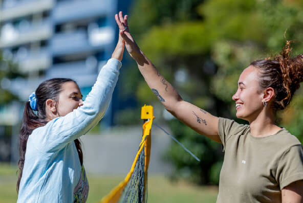 Two people stand on either side of a volleyball net outside in a park. They are both smiling widely and high-fiving each other. 
