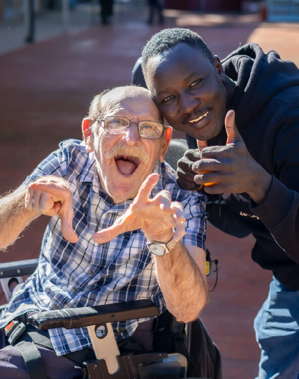 A CPL client sitting in a wheelchair and direct support worker pose for a photo happily. They are cheekily holding their thumbs up for the camera. 