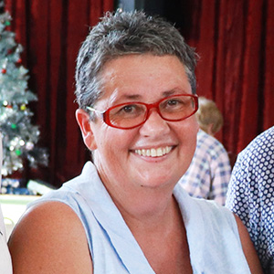 A cropped photo of Felicity wearing a sleeveless blue blouse and red glasses. She has short dark grey hair