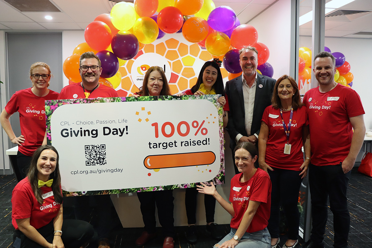 Rhys with a group of people in red giving day t-shirts holding a large sign shoing 100% target reached