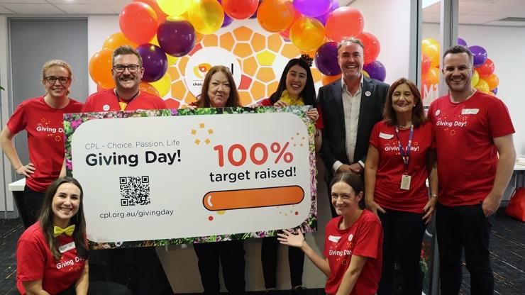 Rhys with a group of people in red giving day t-shirts holding a large sign shoing 100% target reached