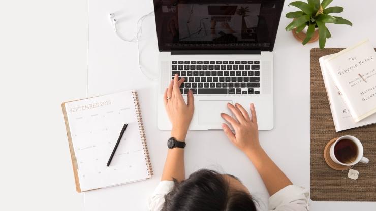 A bird's eye view of a person at a desk. There is a laptop, note pad and pen and a plant.