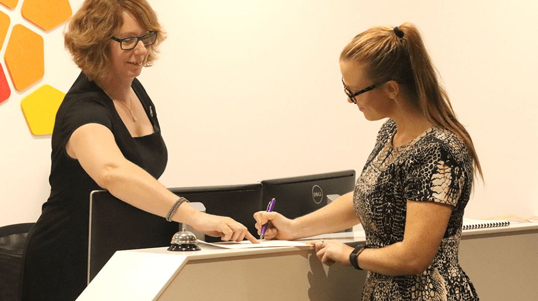 Two woman stand at a reception desk, one signing a piece of paper