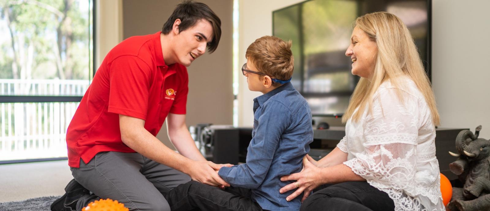 Carter, Daniel and Nicky seated on the loungeroom floor. Daniel is holding a small ball, supported by Carter.